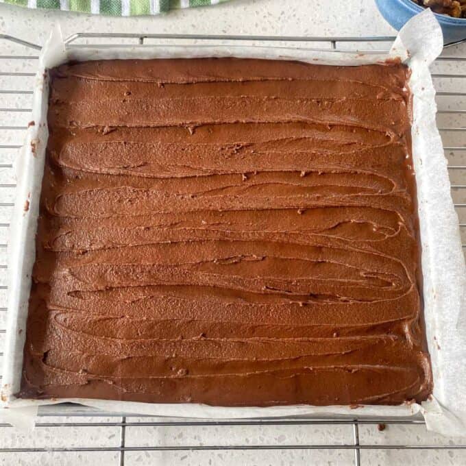 Chocolate Cornflake Slice A square baking pan lined with parchment paper holds a smooth, chocolate-frosted cake cooling on a wire rack.