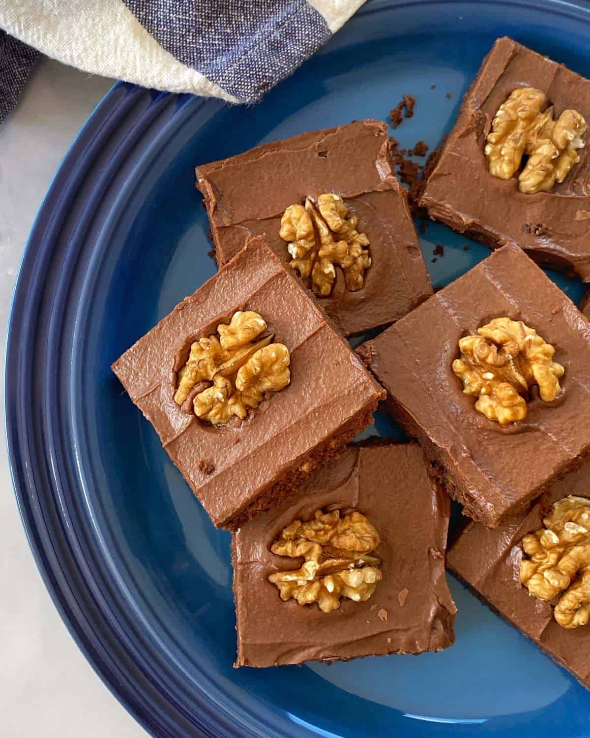 Six chocolate frosted brownies topped with walnut halves are arranged on a blue plate, with a striped napkin partially visible in the corner.
