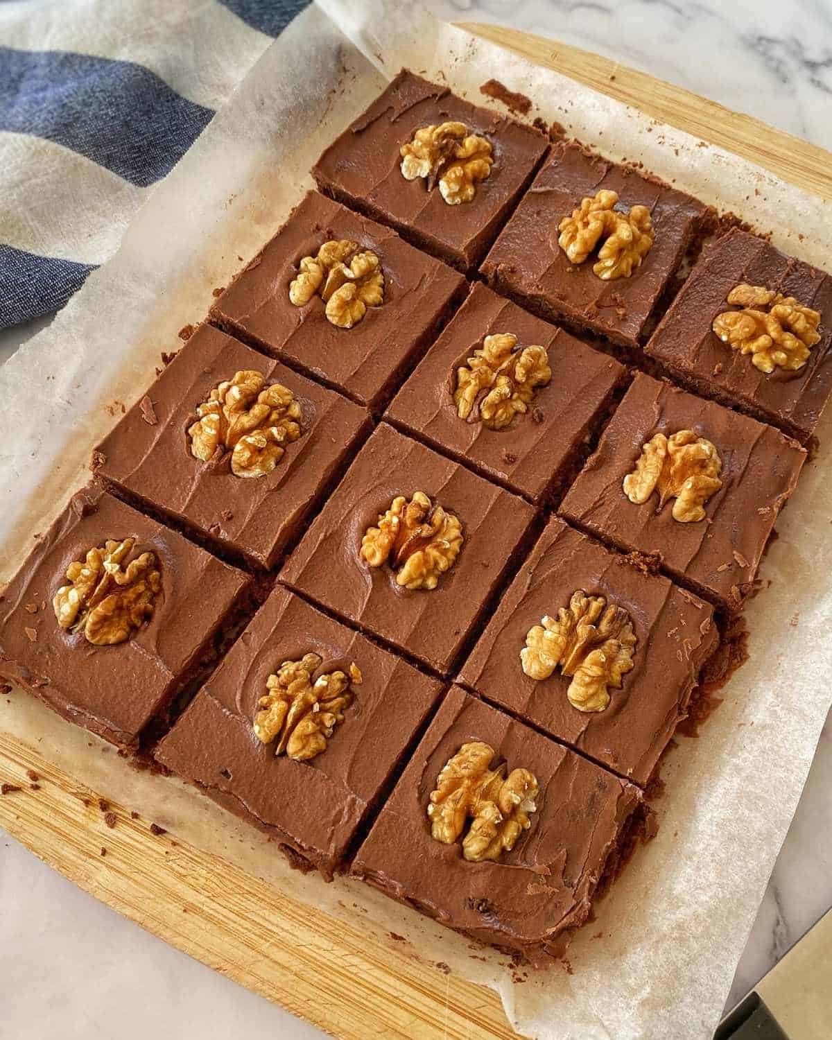 Chocolate Cornflake Slice A tray of chocolate brownies cut into squares, each topped with a walnut half, on parchment paper and a wooden board.