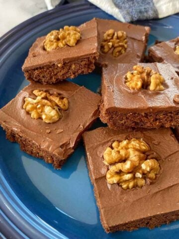 Brownie squares with chocolate frosting, each topped with a walnut half, arranged on a blue plate.