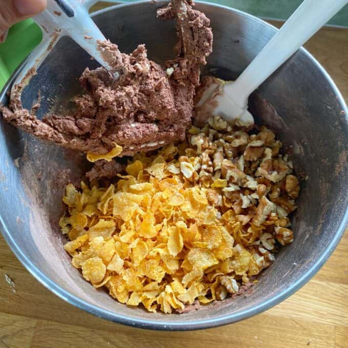 Chocolate Cornflake Slice A metal mixing bowl with chocolate cookie dough, cornflakes, and walnut halves, with two spatulas resting inside.