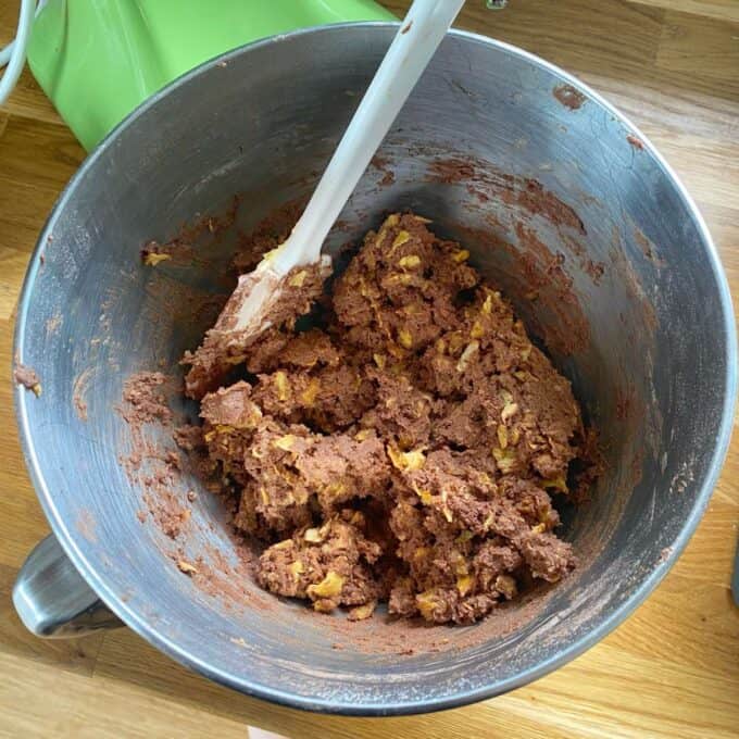 Chocolate Cornflake Slice A metal mixing bowl with chocolate cookie dough and visible chunks, with a white spatula resting inside on a wooden countertop.