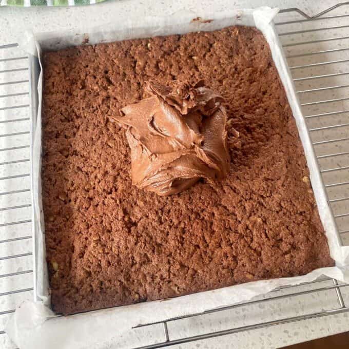 Chocolate Cornflake Slice A square, baked chocolate cake in a lined tin, cooling on a wire rack with a large dollop of chocolate frosting on top.