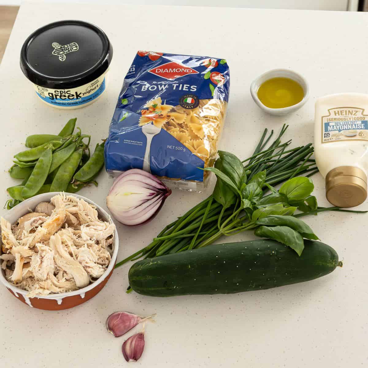 Herby Chicken Pasta Salad Ingredients on a white counter: Greek yogurt, bow tie pasta, olive oil, mayonnaise, snap peas, red onion, chives, basil, shredded chicken, cucumber, mushrooms, and garlic cloves.