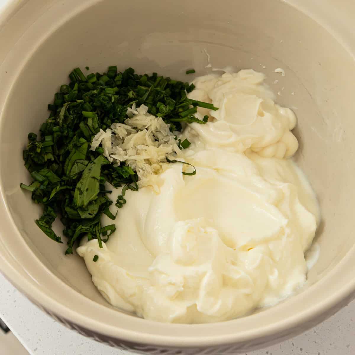 Herby Chicken Pasta Salad A mixing bowl containing mayonnaise, sour cream, chopped green herbs, and minced garlic.