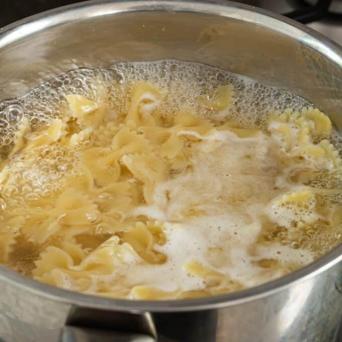 Herby Chicken Pasta Salad A pot of farfalle pasta boiling in water on a stovetop, with foam forming on the surface.