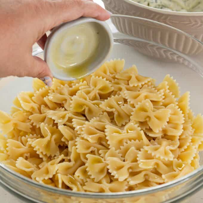 Herby Chicken Pasta Salad A hand pours olive oil from a small bowl onto cooked farfalle pasta in a large glass mixing bowl.