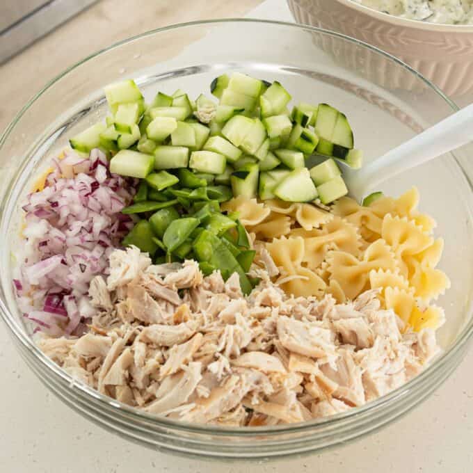 Herby Chicken Pasta Salad A glass bowl containing chopped cucumber, red onion, green bell pepper, cooked bowtie pasta, and shredded chicken, ready to be mixed.