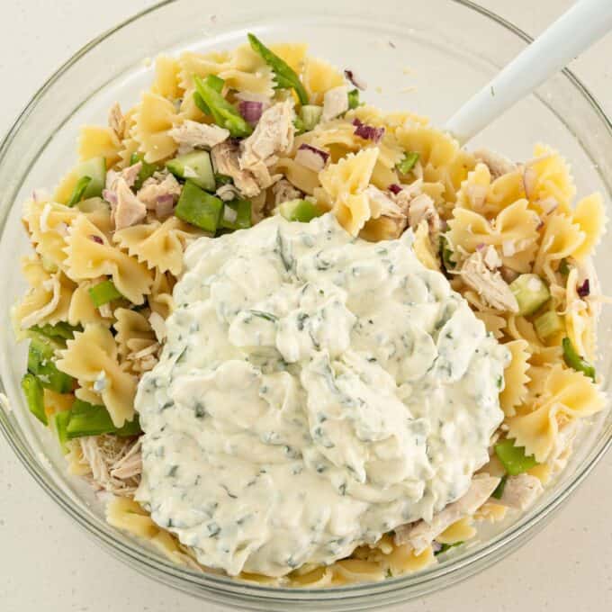 Herby Chicken Pasta Salad A glass bowl filled with bow tie pasta, chopped vegetables, shredded chicken, and a large dollop of creamy herb dressing on top.