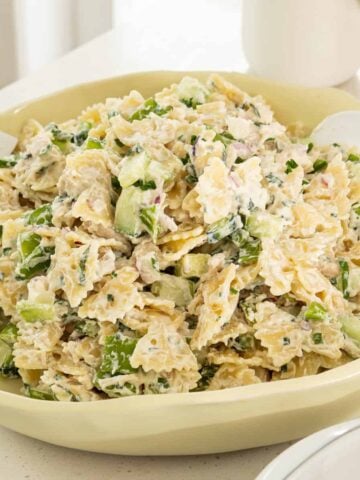 A bowl of pasta salad with bowtie pasta, chopped celery, green onion, and a creamy dressing, served with white salad utensils.