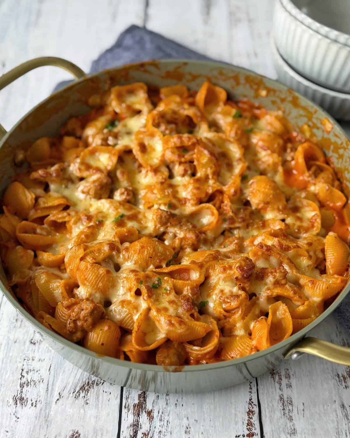Italian Sausage Pasta Bake A skillet filled with baked pasta shells in tomato sauce topped with melted cheese and pieces of meat, set on a rustic white wooden surface.