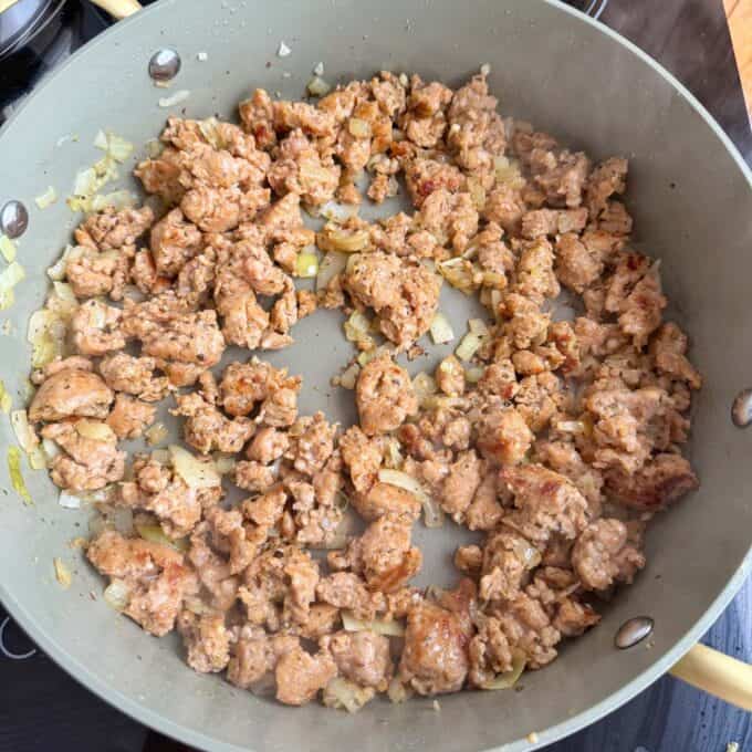 Italian Sausage Pasta Bake Ground meat and diced onions are being cooked and browned in a large skillet on a stovetop.