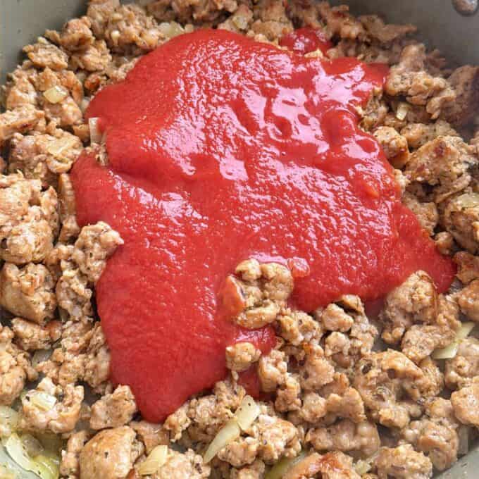 Italian Sausage Pasta Bake Cooked ground meat and onions in a pan topped with a layer of tomato sauce, ready to be mixed.
