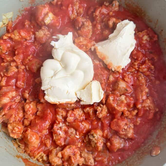 Italian Sausage Pasta Bake Ground meat in tomato sauce with two large portions of cream cheese placed on top in a cooking pot.