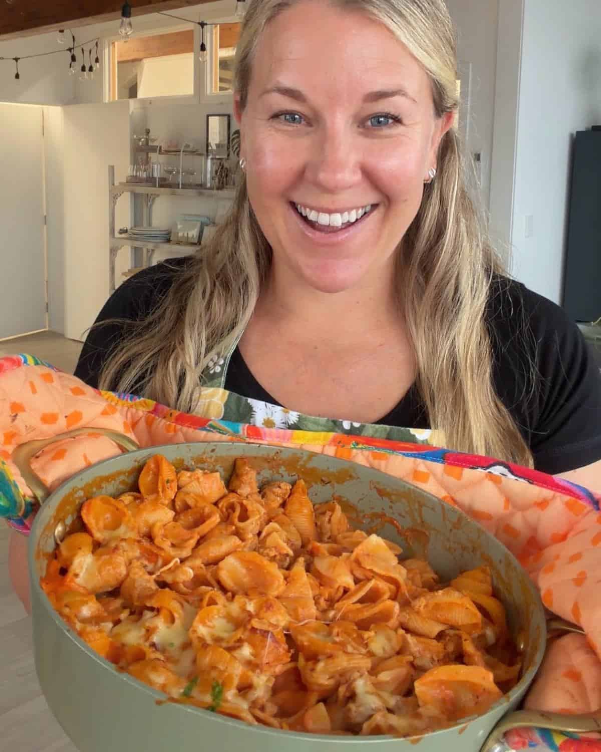 Italian Sausage Pasta Bake A woman smiles while holding a large green dish filled with baked pasta in a kitchen setting.