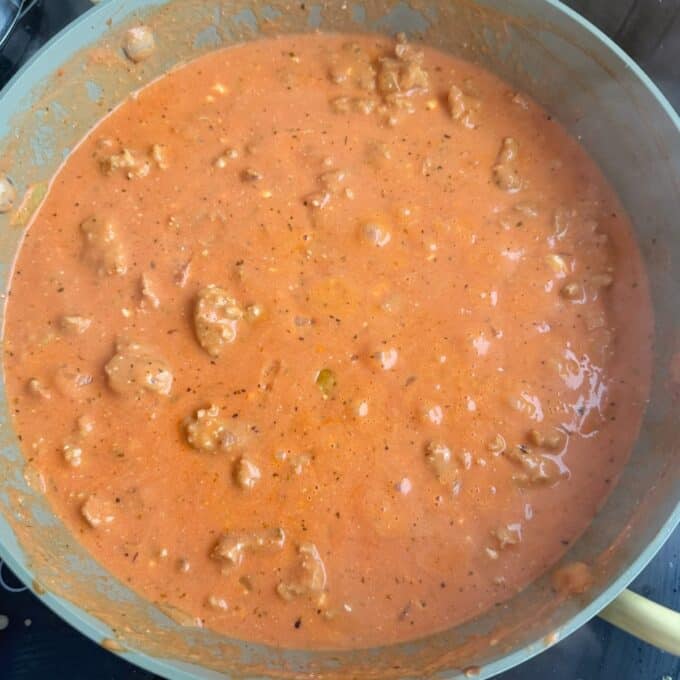 Italian Sausage Pasta Bake A pan filled with creamy tomato-based meat sauce, containing chunks of ground meat and visible herbs, cooking on a stovetop.