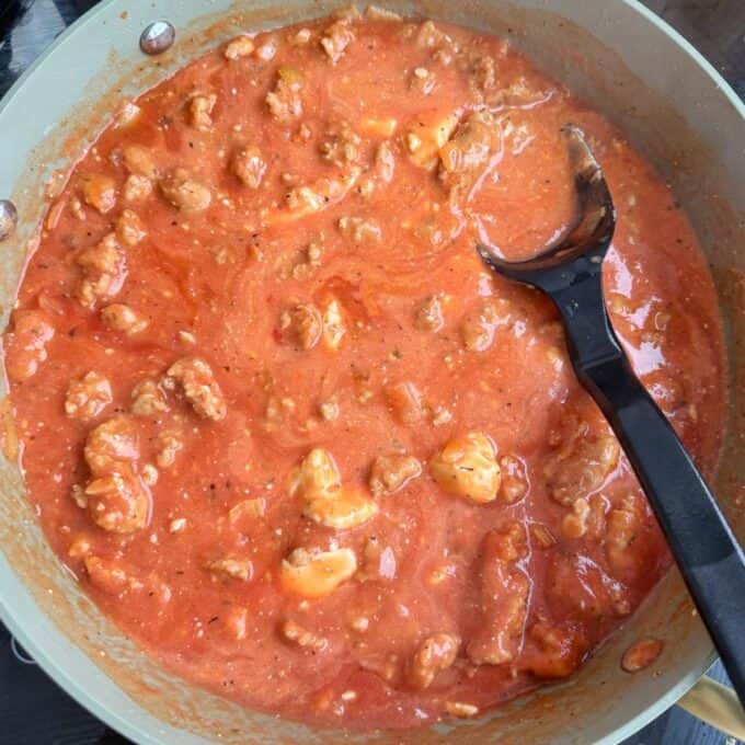 Italian Sausage Pasta Bake A skillet filled with creamy tomato sauce mixed with ground meat and chunks of mozzarella, with a black spoon resting inside.