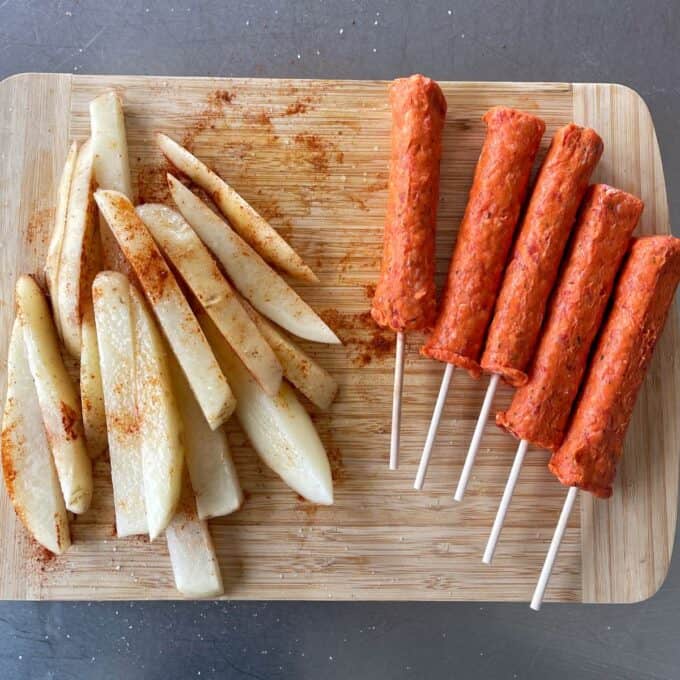 Lamb Koftas on Homemade Flatbreads Raw potato wedges seasoned with spices and five skewered, uncooked kebabs placed on a wooden cutting board.