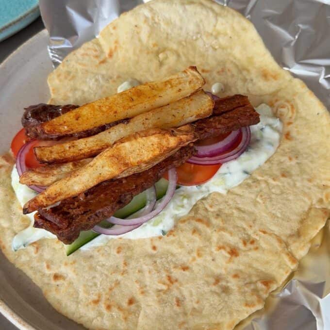 Lamb Koftas on Homemade Flatbreads A flatbread topped with tzatziki sauce, sliced tomato, cucumber, red onion, and fried potato strips on a plate with foil.