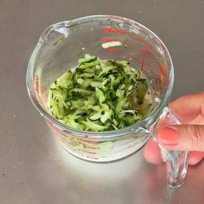 Lamb Koftas on Homemade Flatbreads A hand holding a glass measuring cup filled with shredded cucumber on a gray countertop.