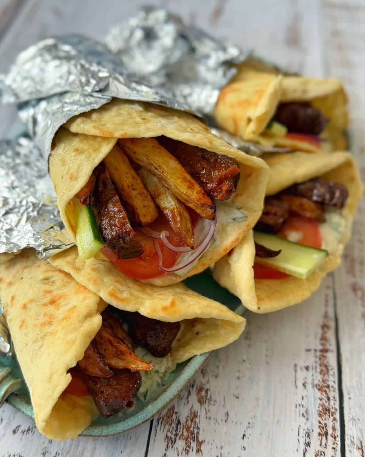 Lamb Koftas on Homemade Flatbreads Three wraps filled with grilled meat, fries, tomato, cucumber, and onions, partially wrapped in foil, placed on a rustic wooden surface.