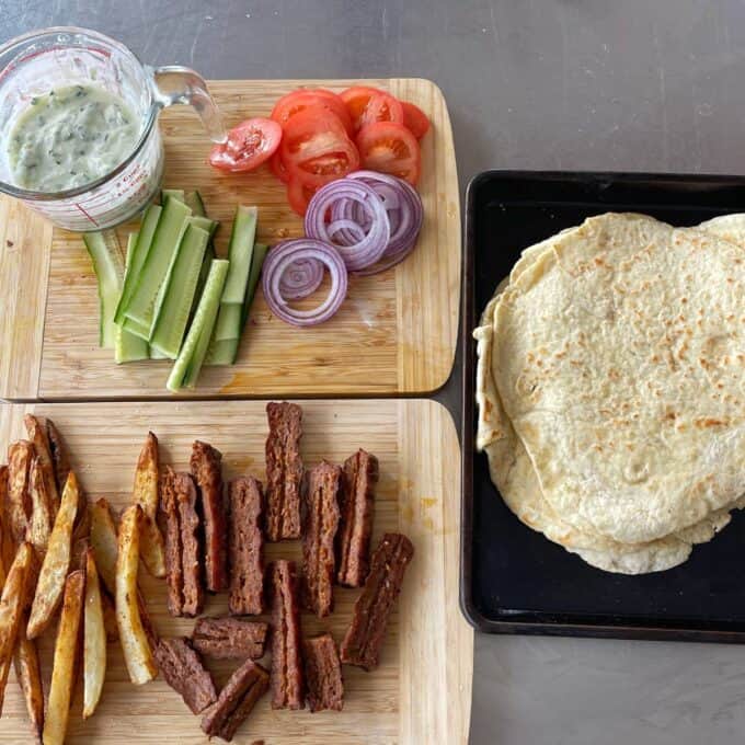 Lamb Koftas on Homemade Flatbreads Chopped tomatoes, red onion, cucumber, and tzatziki on one board; fries and sliced seitan on another; stack of flatbreads on a tray.