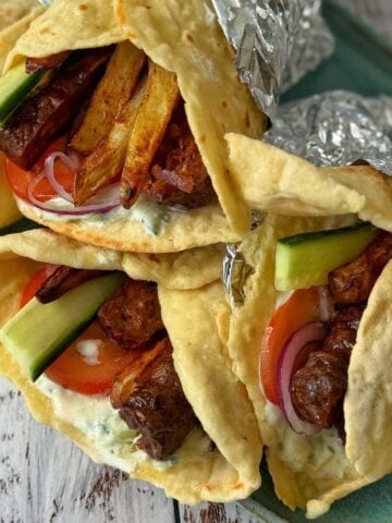 Three wrapped gyros filled with grilled meat-like lamb koftas on homemade flatbreads-tomato, cucumber, onion, fries, and tzatziki sauce, arranged on a tray with foil partially covering the pita.