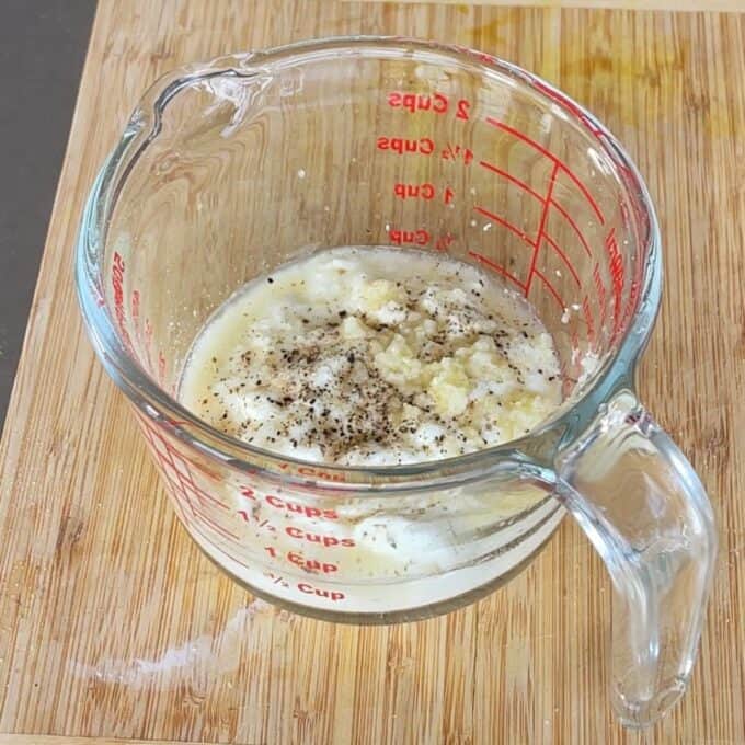 Lamb Koftas on Homemade Flatbreads A glass measuring cup on a wooden board contains a mixture of white liquid, minced garlic, and ground black pepper.