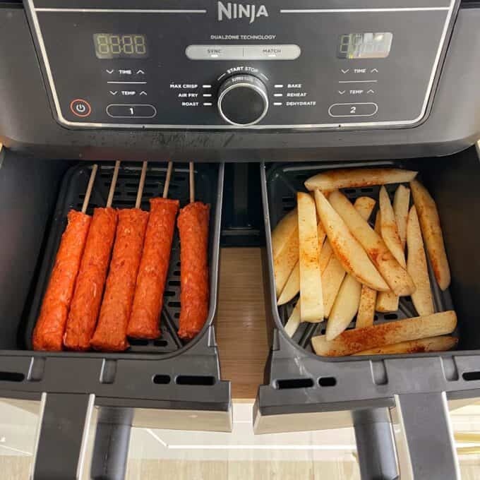 Lamb Koftas on Homemade Flatbreads An air fryer with two compartments; one side contains raw kebab skewers, and the other side contains seasoned potato wedges, both ready to be cooked.