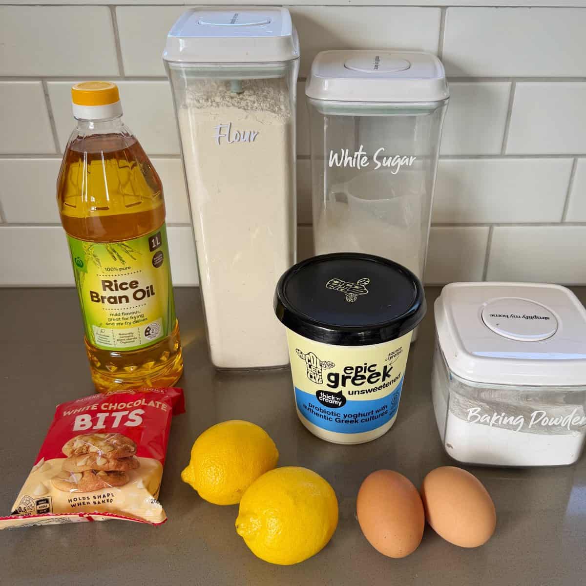 Lemon, Yoghurt and White Chocolate Muffins A collection of baking ingredients on a counter: rice bran oil, flour, white sugar, Greek yogurt, baking powder, chocolate bits, two lemons, and two eggs.