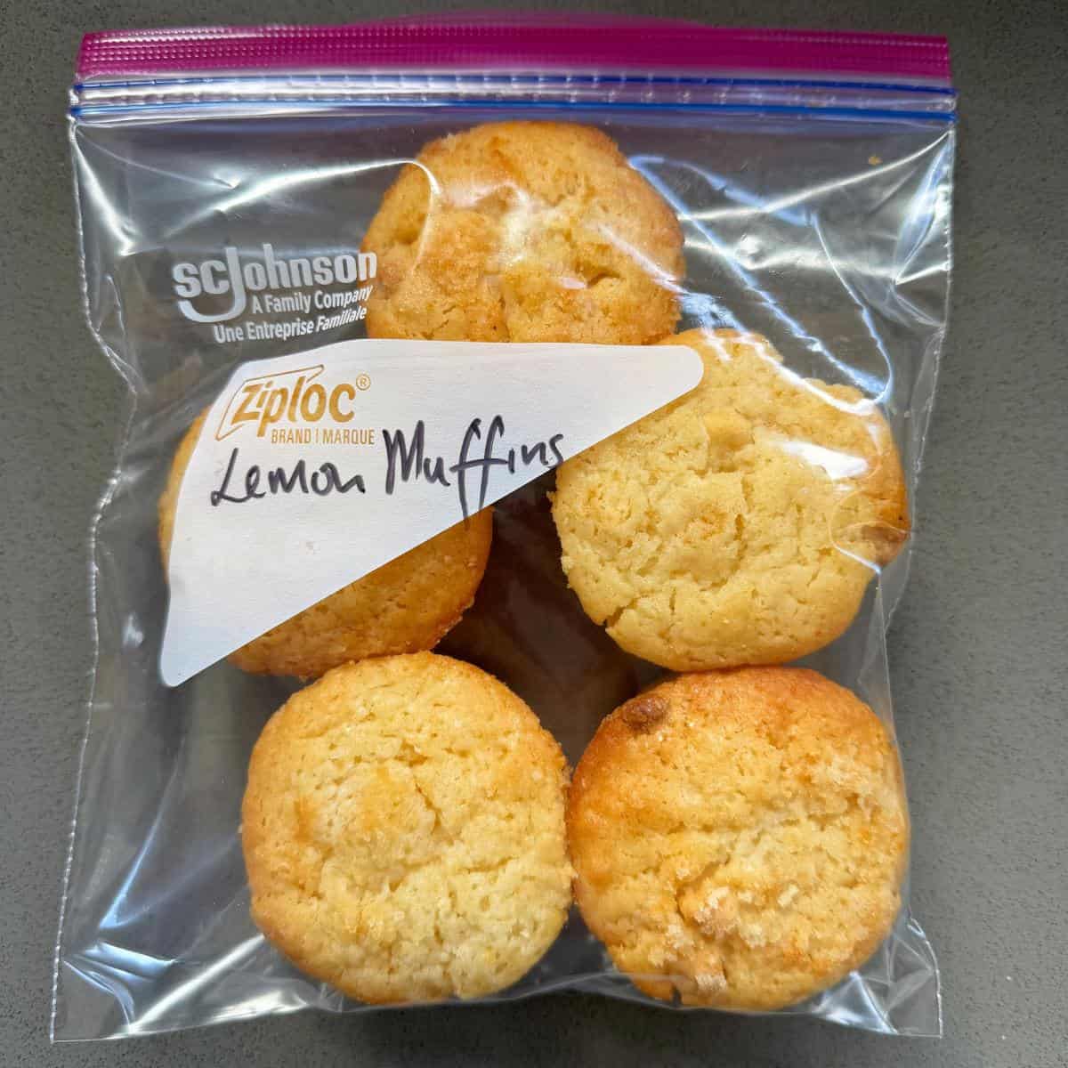 A clear Ziploc bag labeled Lemon Muffins contains five small, golden-brown muffins on a gray surface.