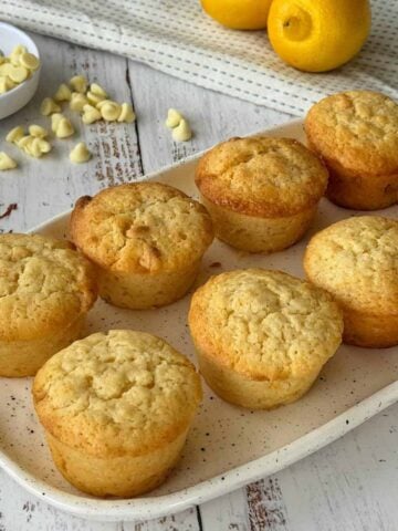 Eight lemon muffins are arranged on a rectangular plate, with white chocolate chips and whole lemons visible in the background on a rustic wooden table.