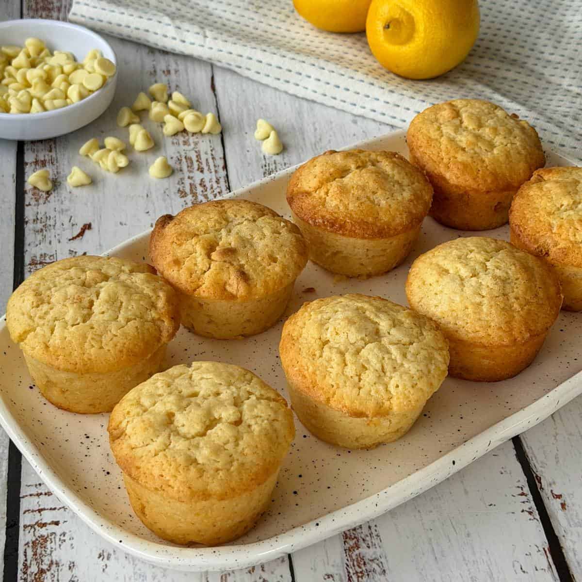 Lemon, Yoghurt and White Chocolate Muffins Eight lemon muffins are arranged on a rectangular plate, with white chocolate chips and whole lemons visible in the background on a rustic wooden table.
