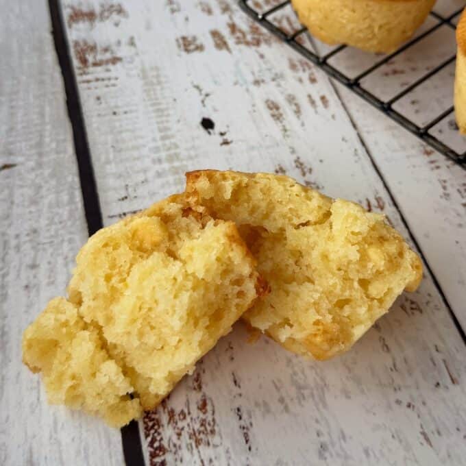 Lemon, Yoghurt and White Chocolate Muffins A split corn muffin sits on a rustic white wooden surface, showing its moist, crumbly interior; more muffins are on a cooling rack in the background.