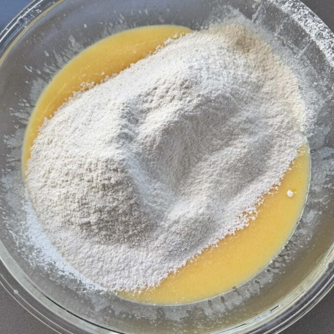 Lemon, Yoghurt and White Chocolate Muffins A glass bowl containing a mound of flour on top of a yellow liquid batter, ready to be mixed.