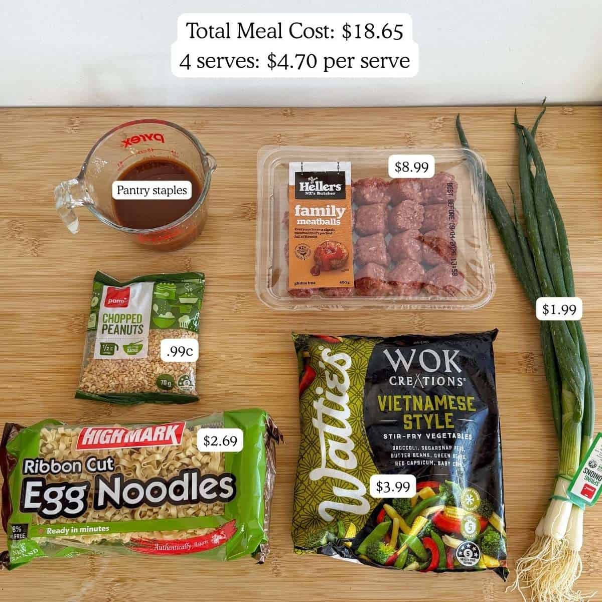 Ingredients for a meal are displayed on a table: sauce in a measuring cup, chopped peanuts, meatballs, green onions, egg noodles, and a bag of Vietnamese-style vegetables.