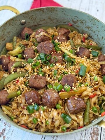 A pan filled with noodles, meatballs, baby corn, snap peas, broccoli, and chopped green onions, garnished with sesame seeds on a wooden surface.