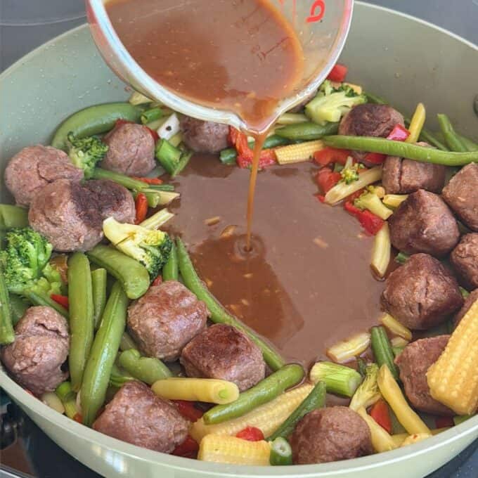 Meatball Noodle Stir Fry A measuring cup pours brown sauce into a pan with meatballs and mixed vegetables including broccoli, sugar snap peas, baby corn, and red bell pepper.