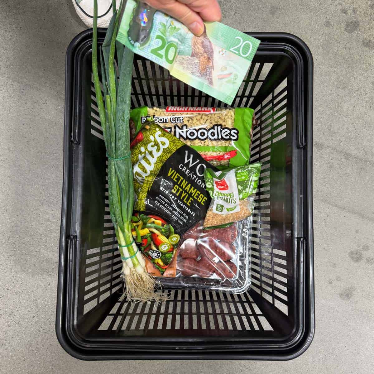 Meatball Noodle Stir Fry A hand holds a 20-dollar bill above a shopping basket containing green onions, noodles, mixed vegetables, chopped peanuts, and packaged beef.