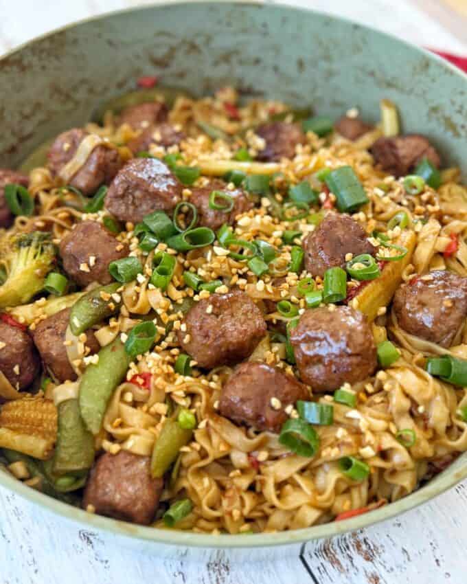 Meatball Noodle Stir Fry A skillet filled with noodle stir fry, meatballs, baby corn, sliced vegetables, chopped green onions, and a sprinkle of sesame seeds.