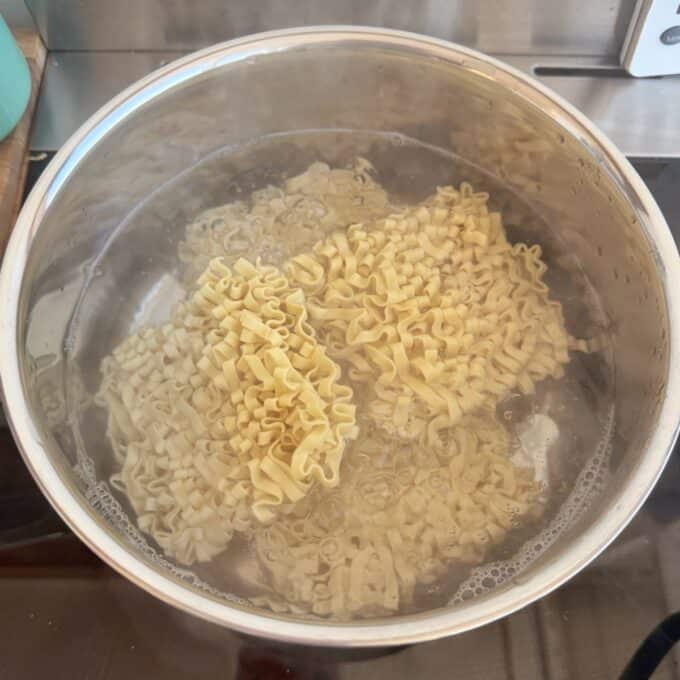 Meatball Noodle Stir Fry A pot of water on a stove with uncooked instant ramen noodles being boiled.