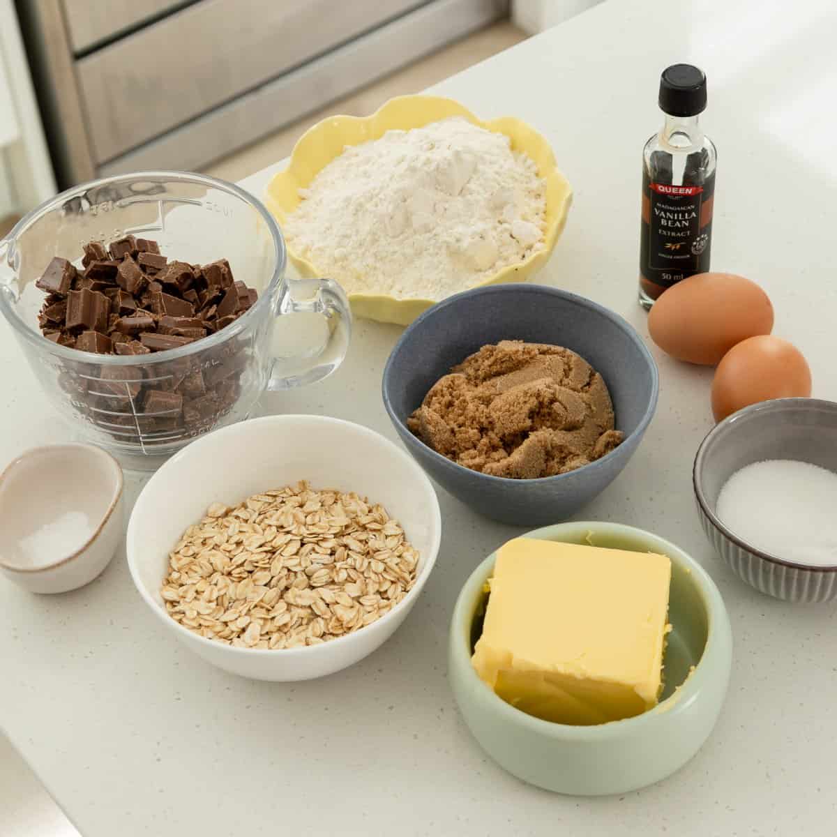 Various baking ingredients on a kitchen counter, including flour, chocolate pieces, oats, butter, brown sugar, white sugar, eggs, and a bottle of vanilla extract.