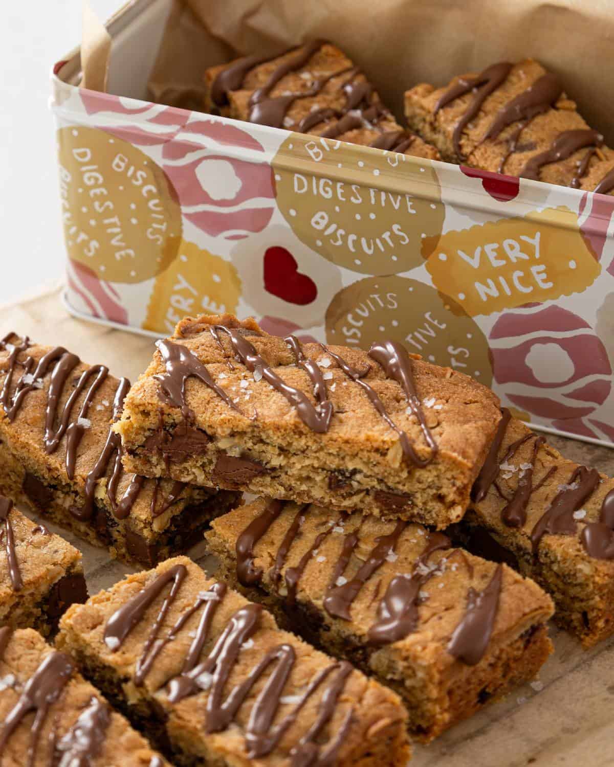 Chocolate-drizzled biscuit bars are arranged on a wooden surface, with more stacked in a patterned tin labeled Digestive Biscuits and Very Nice.