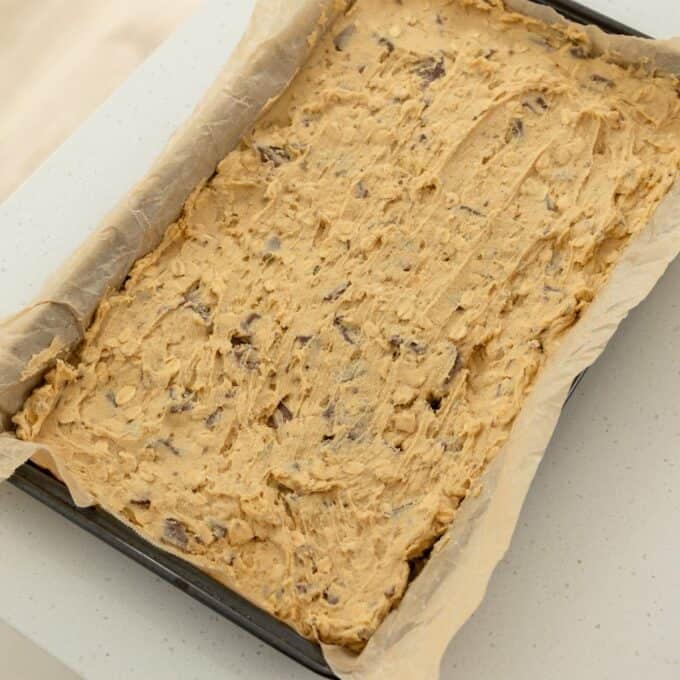 Oaty Choc Chunk Cookie Bars A baking tray lined with parchment paper filled with unbaked cookie dough, spread evenly and ready to go into the oven.