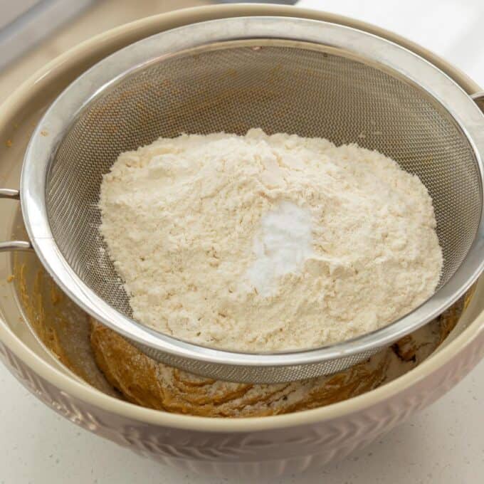 Oaty Choc Chunk Cookie Bars A metal sieve with flour and baking soda rests over a mixing bowl containing a dough mixture.