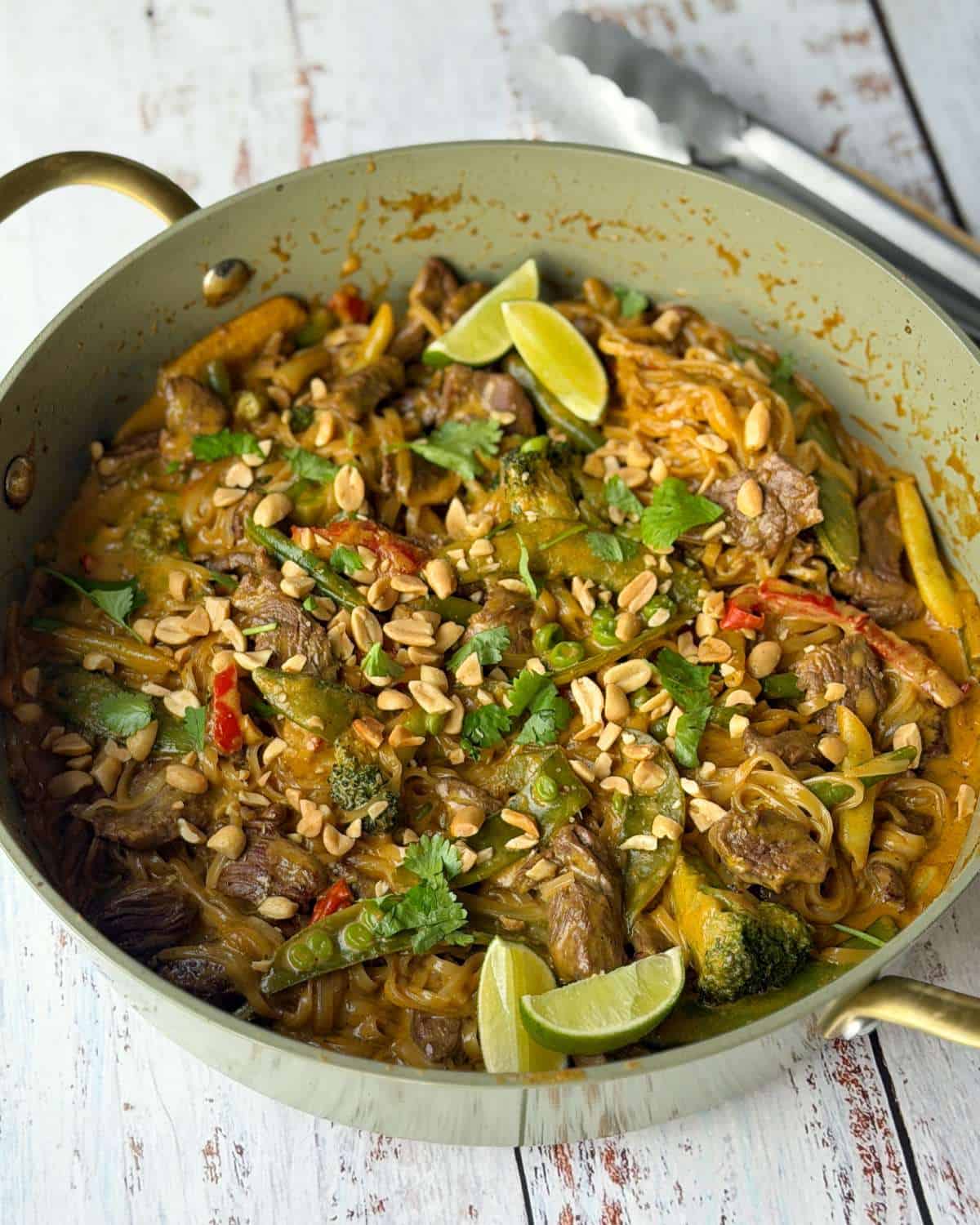 A pan of beef and noodle stir-fry with vegetables, peanuts, cilantro, and lime wedges.