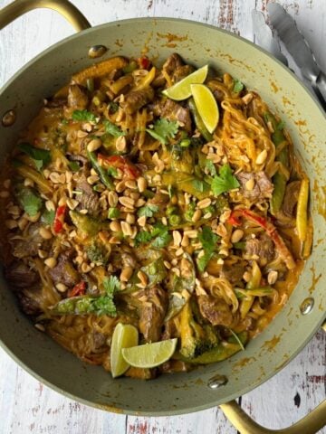 A green pan filled with beef and vegetable stir-fry with noodles, topped with chopped peanuts and lime wedges.