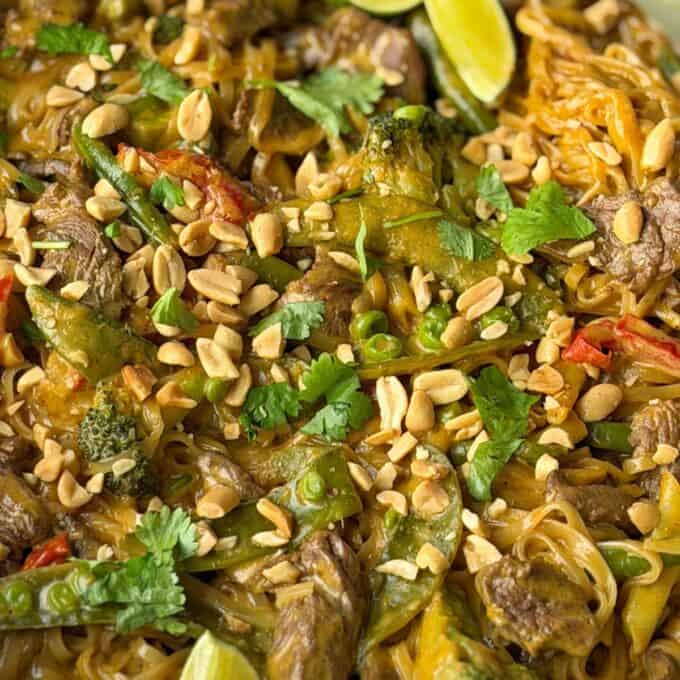 One Pot Beef Curry with Noodles Close-up of a noodle dish with beef, broccoli, snap peas, peanuts, and cilantro, garnished with lime wedges.