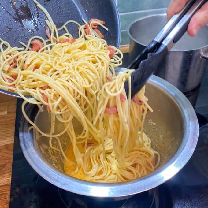 Spaghetti Carbonara Cooked spaghetti with pieces of bacon is being transferred from a pan into a metal bowl using tongs, with a pot visible in the background.