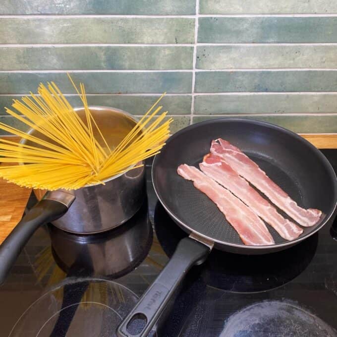 Spaghetti Carbonara Uncooked spaghetti is sticking out of a pot of water on a stove next to a frying pan with raw bacon strips.